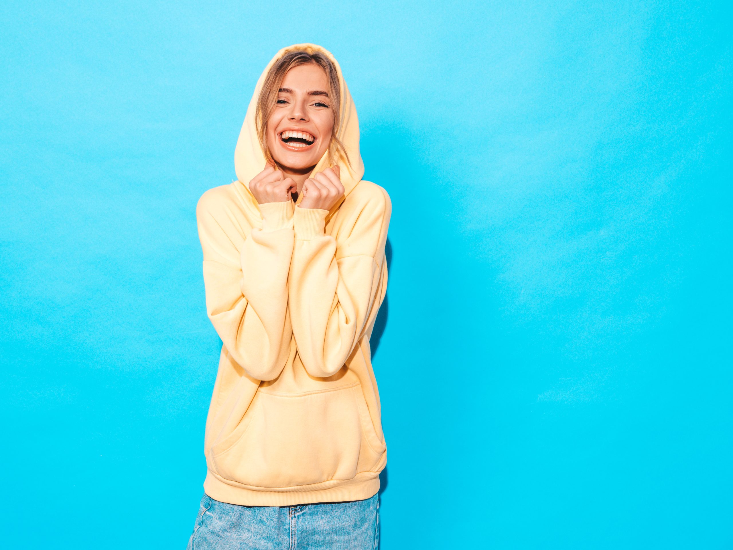 Portrait of young beautiful smiling girl in trendy summer hipster yellow hoodie.carefree woman posing near blue wall in studio. Positive model having fun
