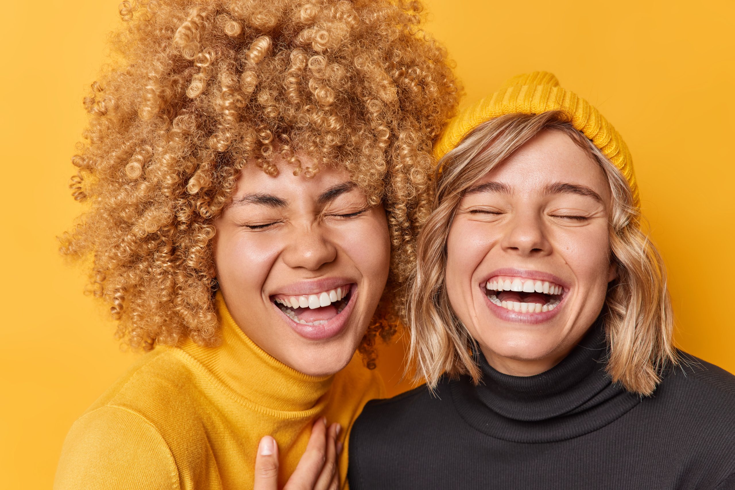 Portrait of two female friends have fun laugh joyfully keep eyes closed smile toothily dont hide sincere feelings dressed in casual turtlenecks isolated over yellow background. People and emotions