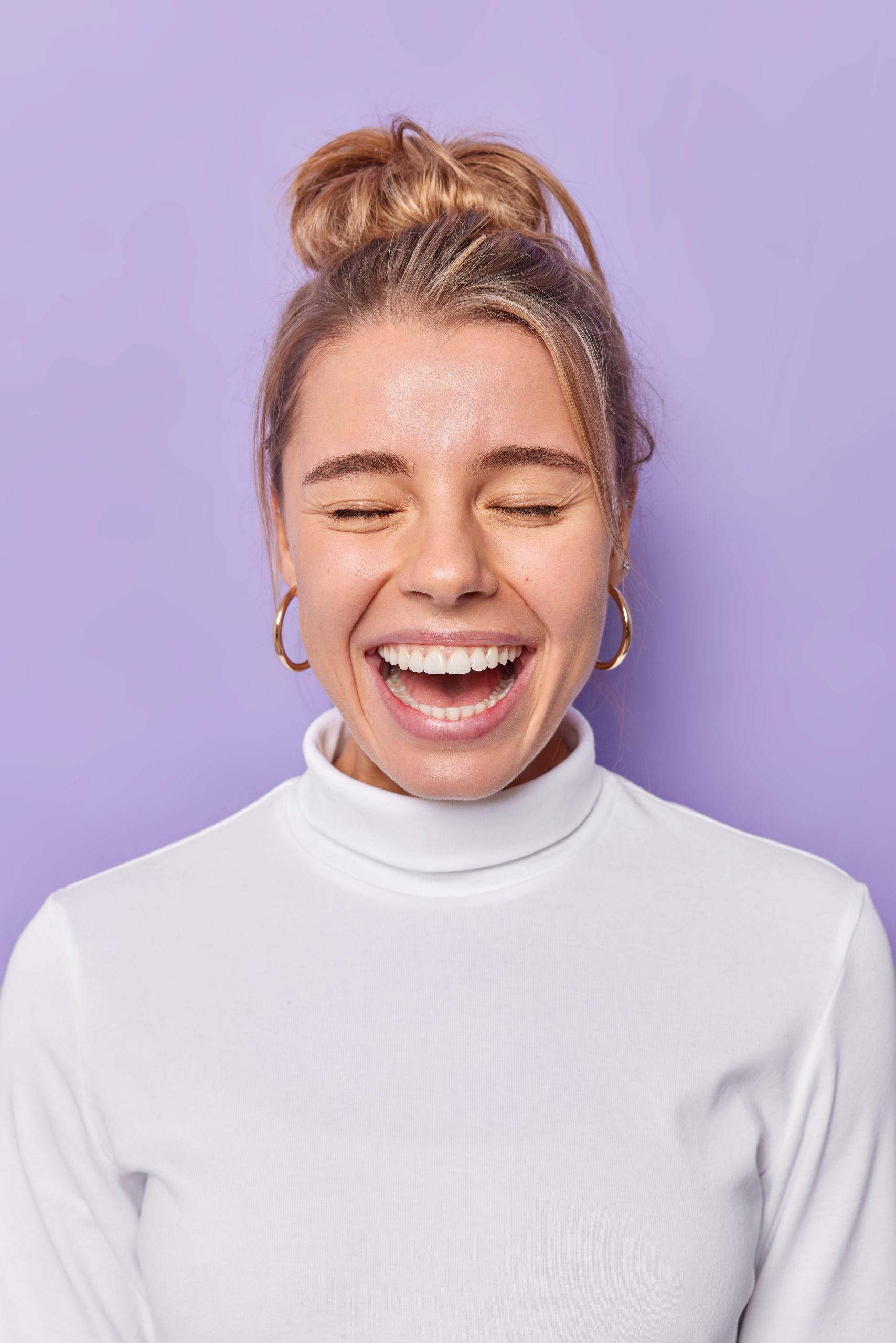 Joyful young woman with healthy skin laughs happily feels very glad keeps eyes closed dressed in casual turtleneck isolated over purple background. People positive emotions and feelings concept